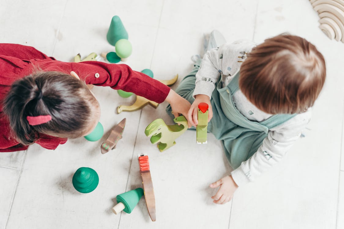 Two young children playing with wooden dinosaur toys and green shapes on a white floor